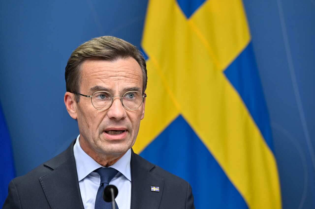 A man wearing a suit, tie, and glasses. He is standing at a lectern and speaking in front of a microphone. The Swedish flag is behind him.
