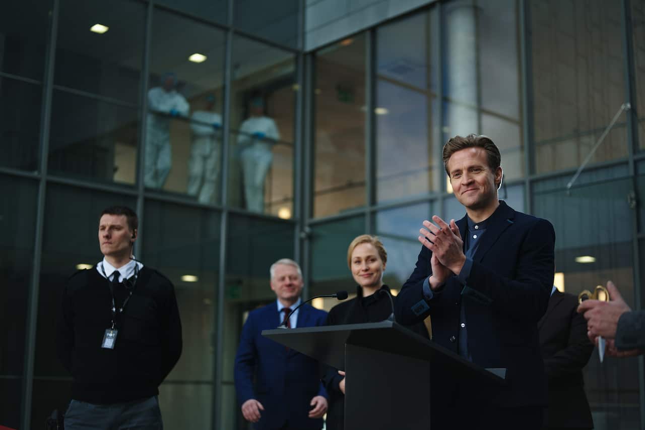 A man in a suit stands at a lectern, applauding. Several people stand behind him, with approving expressions. 