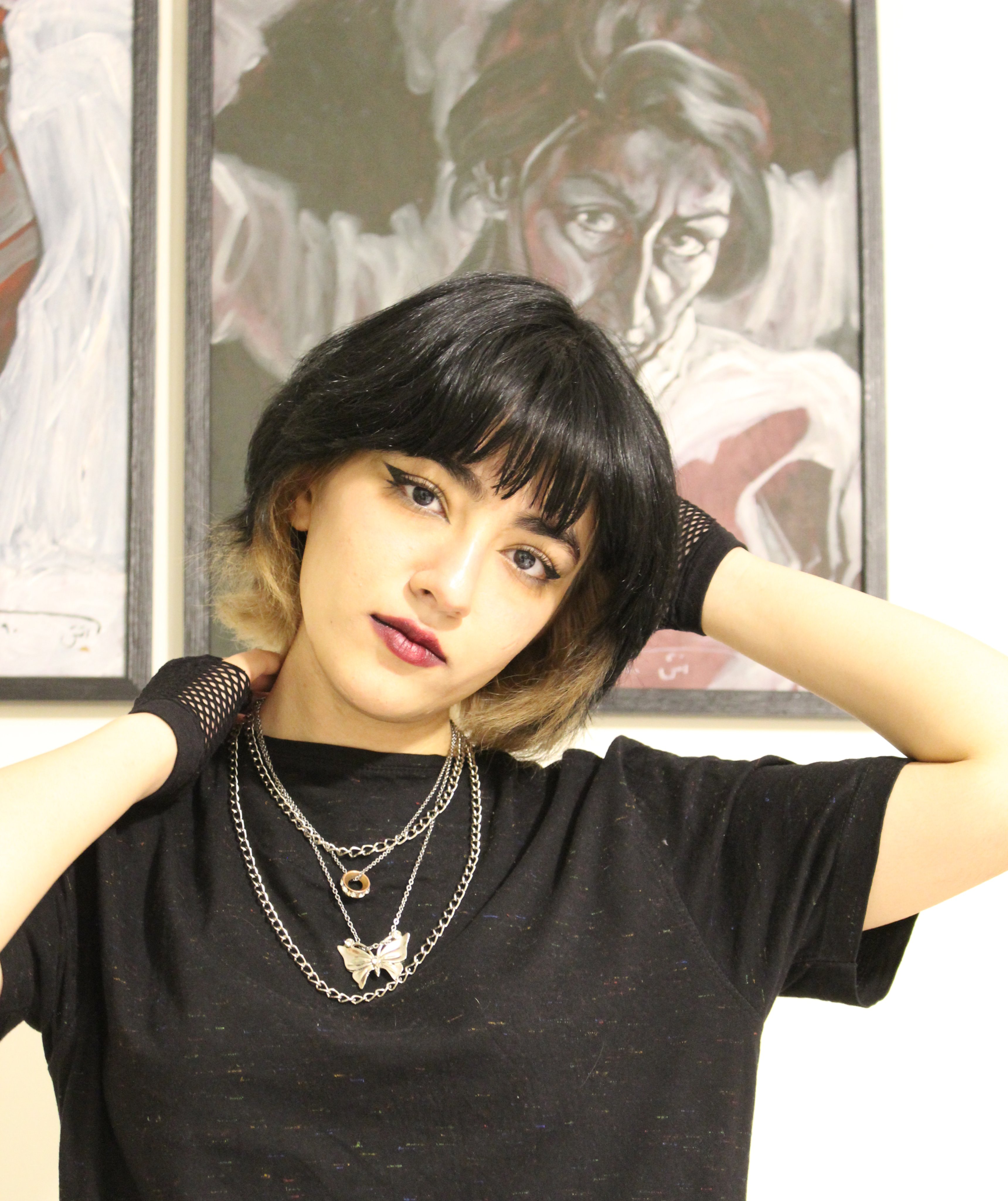 A girl with short hair poses in a black t-shirt and is wearing layered necklaces. 