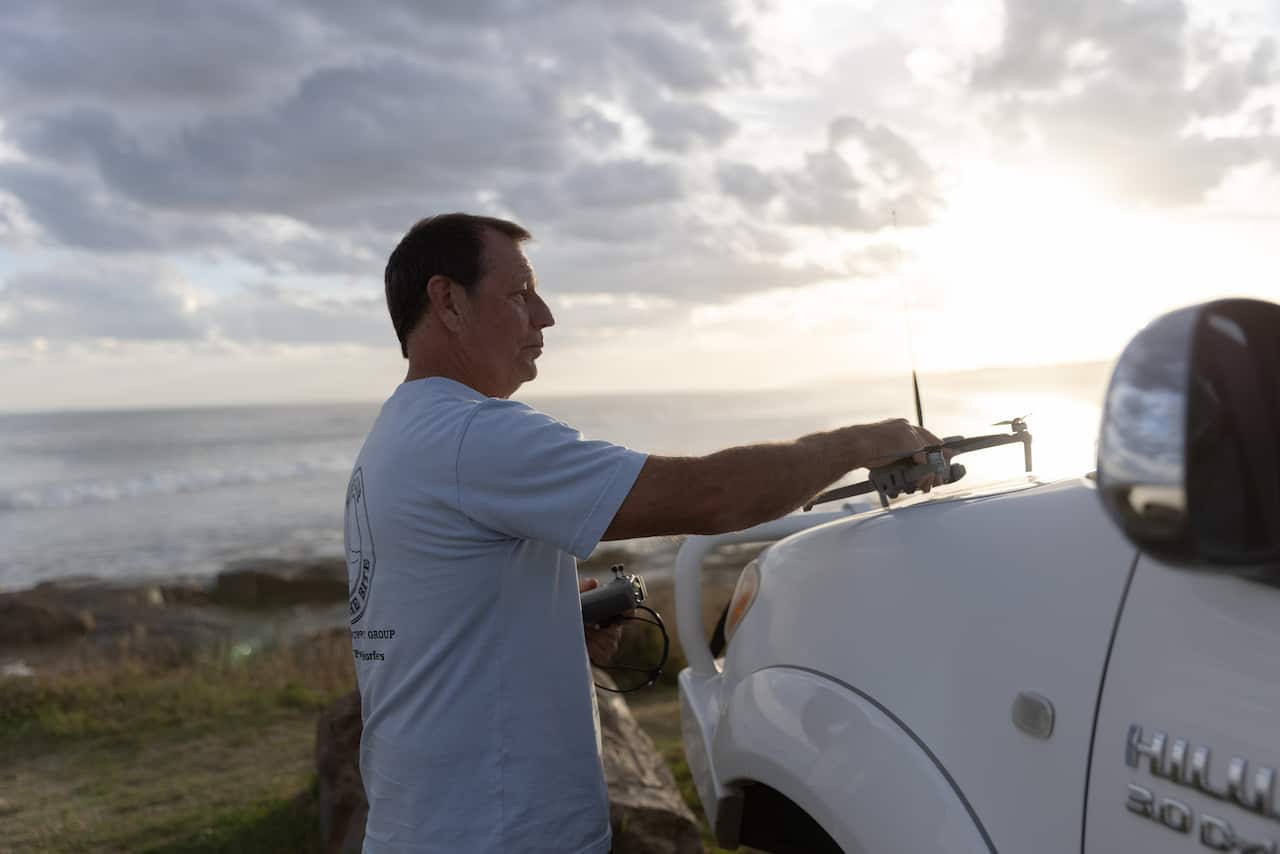 A man sits a drone on top of his car, which is overlooking the beach. 