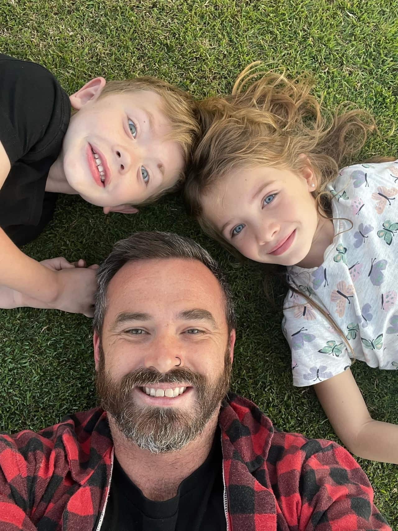 A dad with his two young children lying on the grass together. They are smiling.