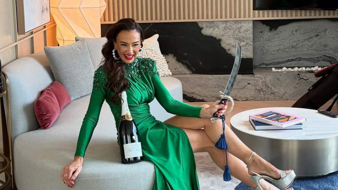 a woman in a green sequinned dress and silver heels sits on a couch with a bottle of champagne and a sabre