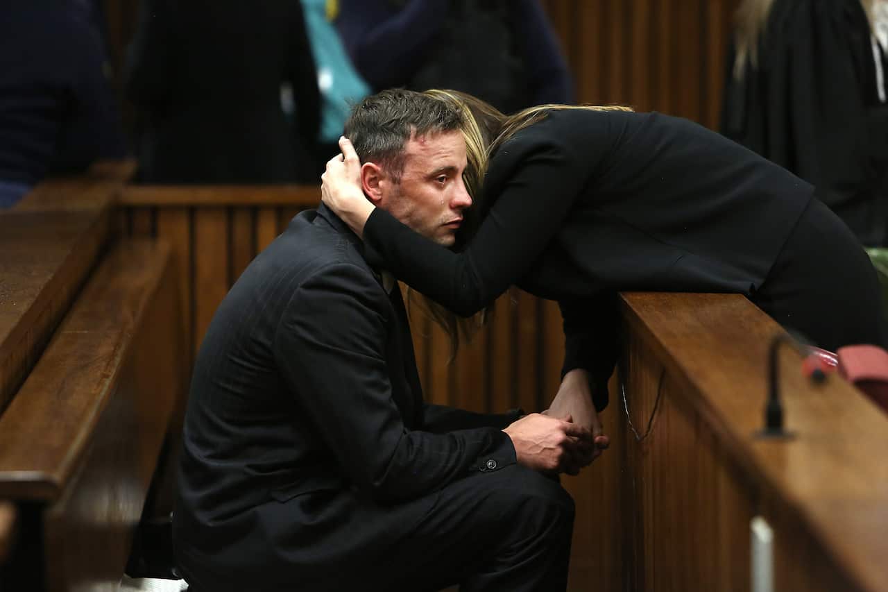 A man sits in court as a woman leans over and hugs him.