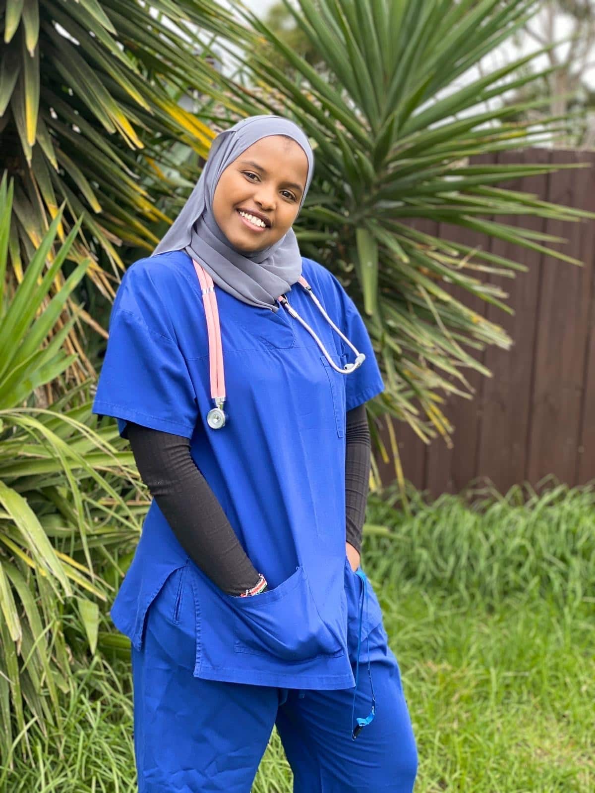 A smiling healthcare professional wearing a grey hijab, blue scrubs, and a stethoscope stands outdoors in front of lush green plants.