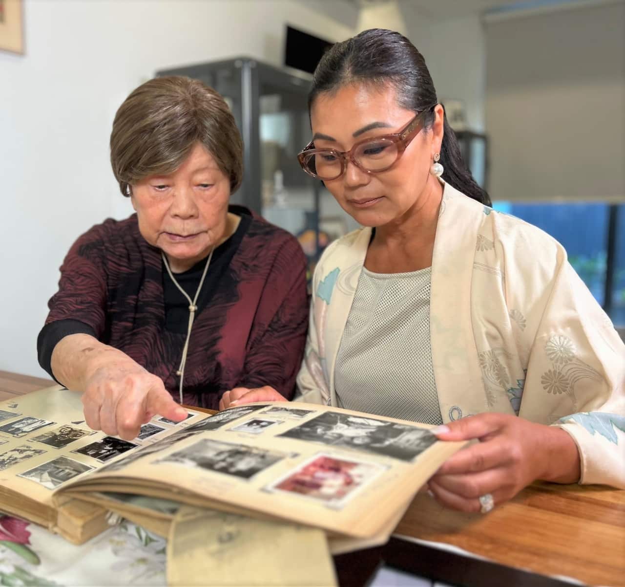 An elderly woman in a burgundy top sits at a table looking at old photos with a younger woman in a pale kimono.