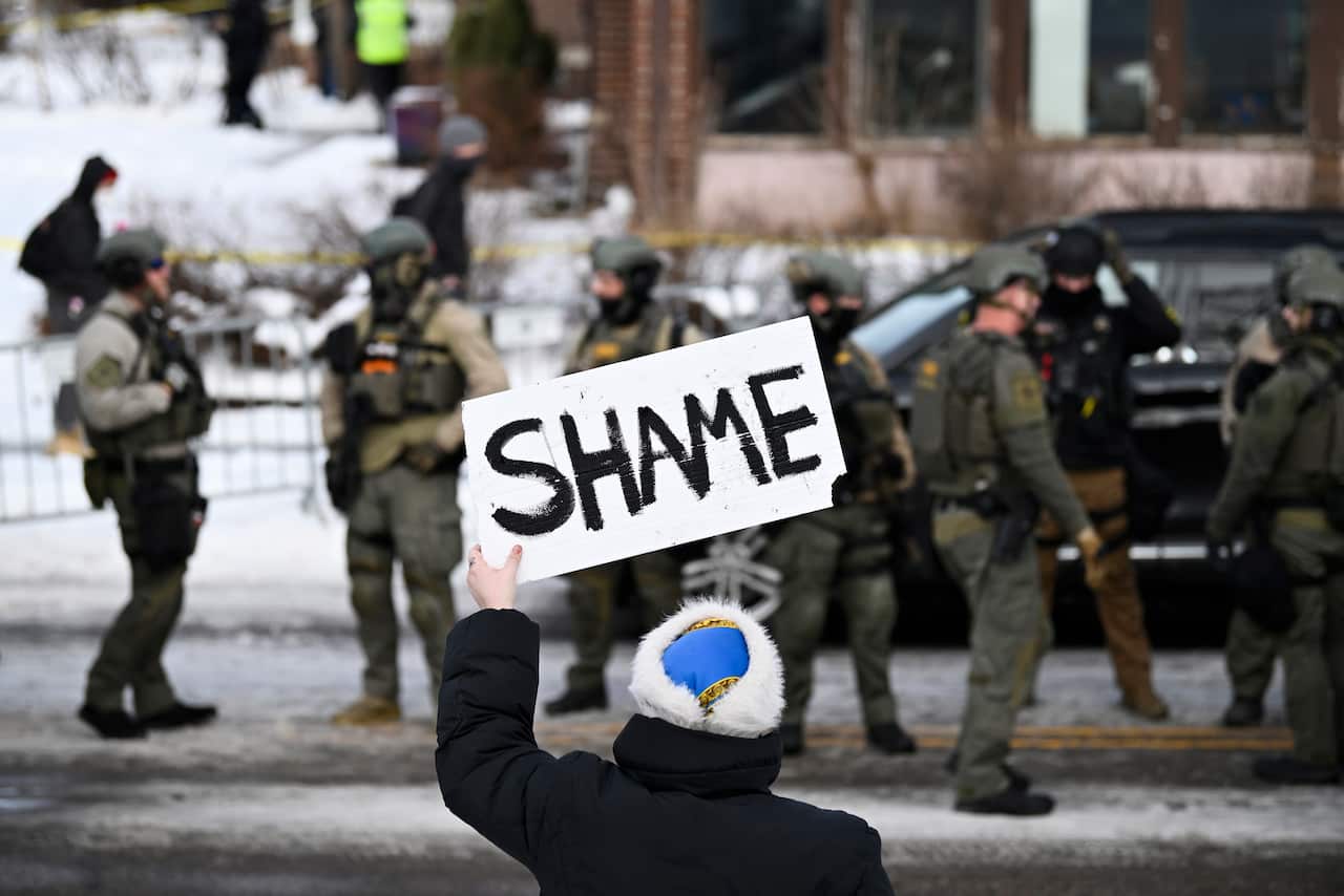 An onlooker holds up a sign that reads "shame" in front of several law enforcement agents.