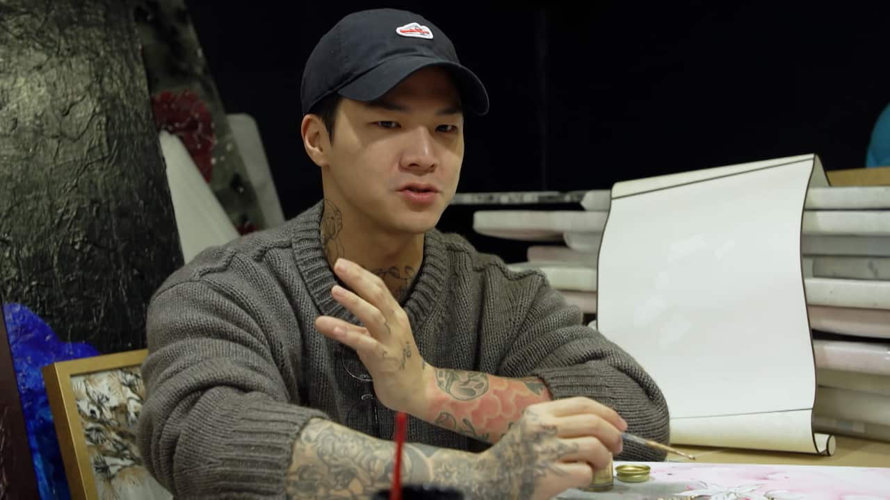 Min Su wearing a thick grey sweater and black baseball cap paints with a brush in his right arm.