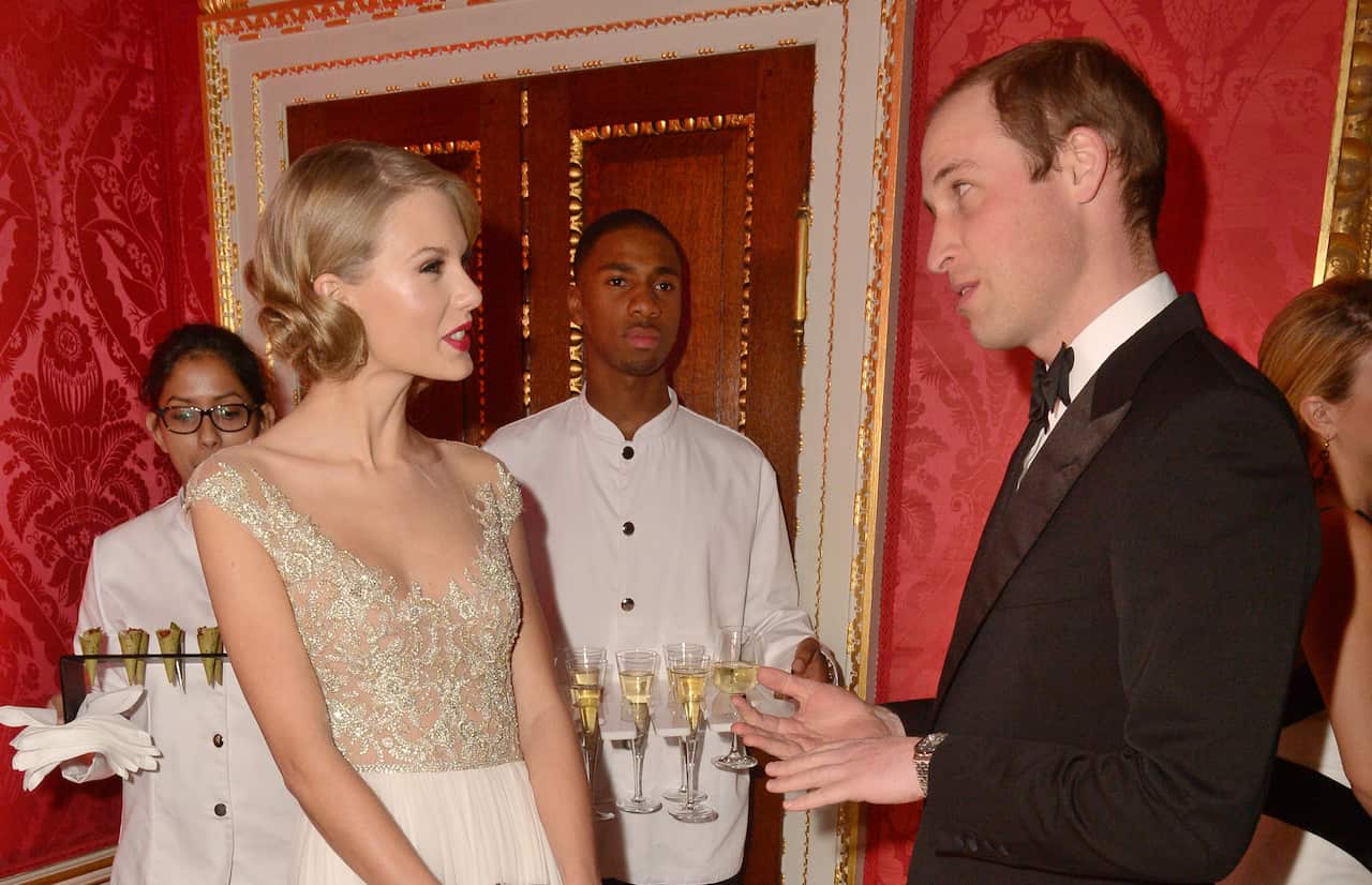 A woman in a white and gold dress with blonde hair talks to a prince in a suit.