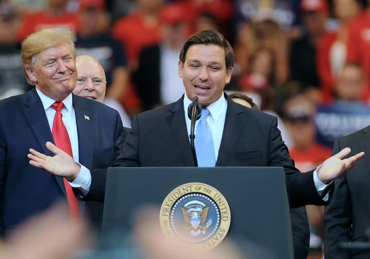 Ron DeSantis speaks at a rally. Donald Trump looks at him with a smile. 