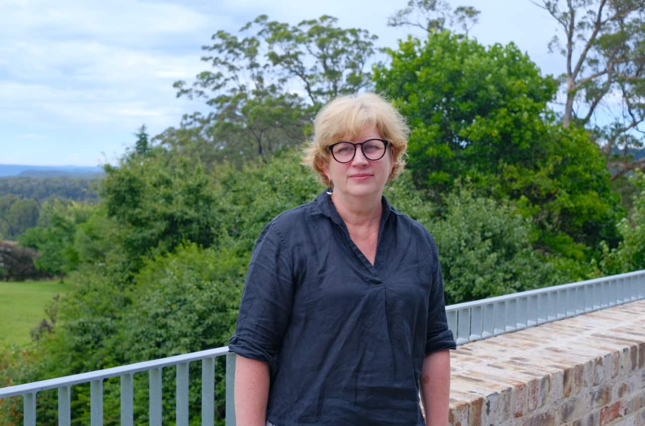 A woman in a dark blouse stands in front of a garden