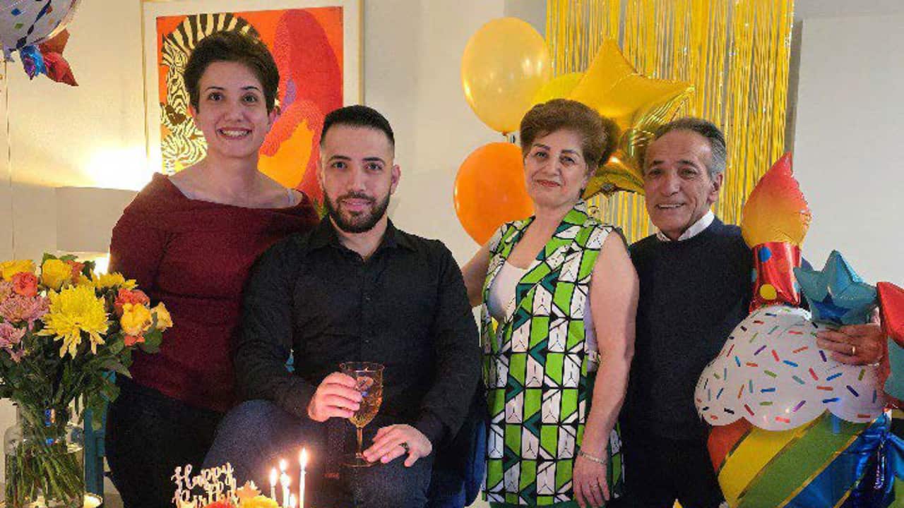Four people pose together at a brightly decorated birthday celebration, with a lit cake, balloons and flowers in the foreground and festive streamers behind them.