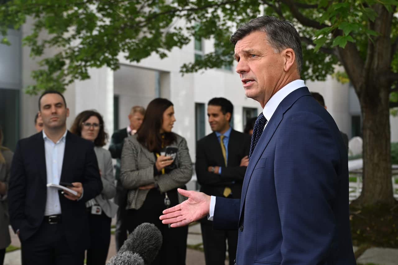 Angus Taylor, wearing a suit, speaking outside before microphones. A group of journalists is standing in the background