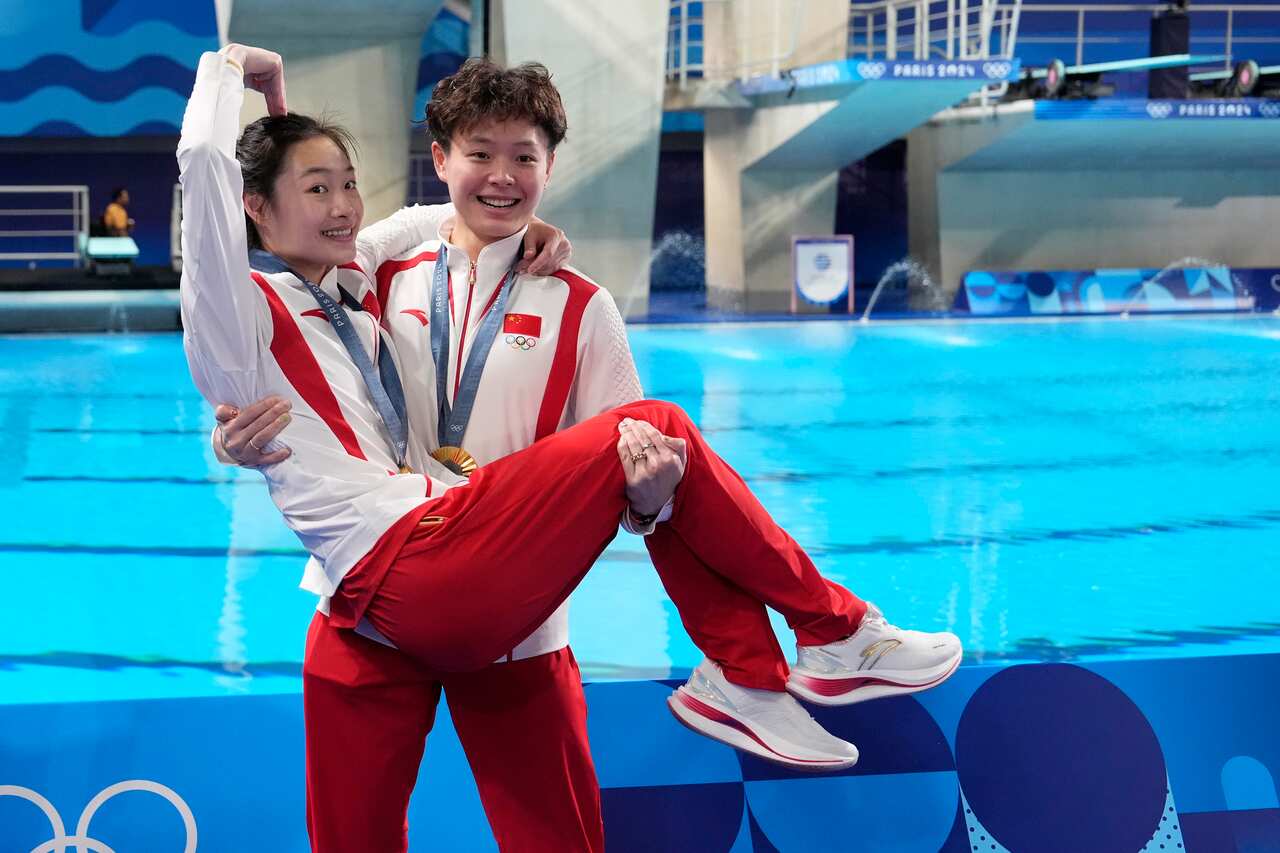Two women in tracksuits of red bottoms and white tops. One is carrying the other in front of a diving pool.
