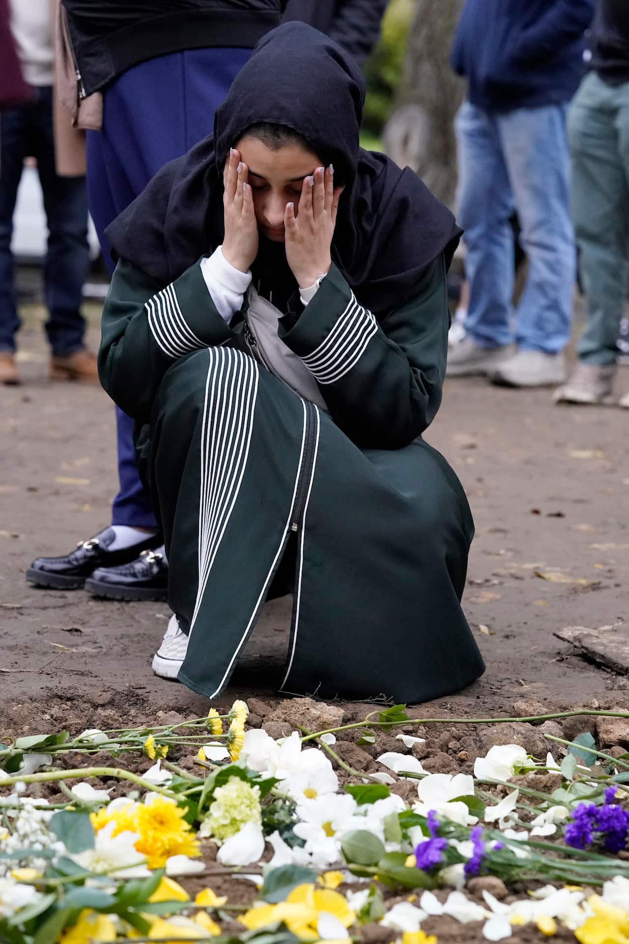 A women wearing dark-coloured clothes kneels in front of a grave with her head in her hands.