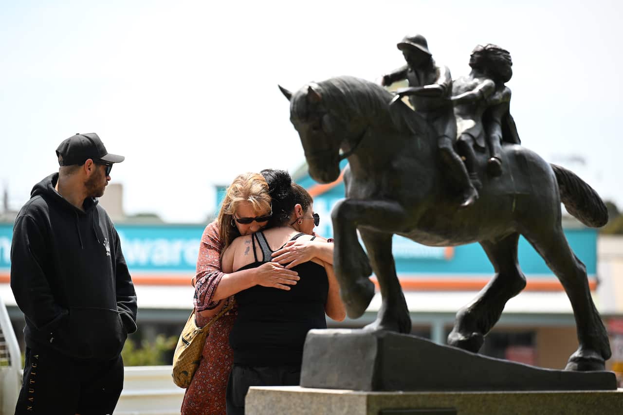 A person in a black hoodie stands next to two women emotionally embracing each other, in front of a statue of a horse with two children.