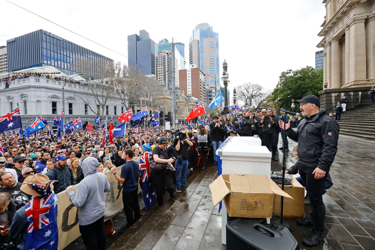 A man speaking on the steps of a building to a large crowd of people, many holding Australian flags.