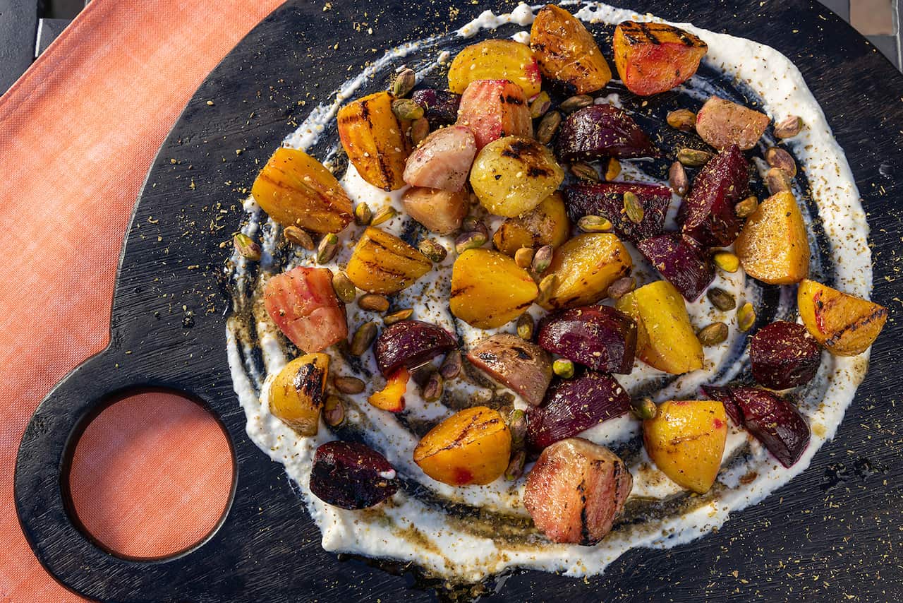 On a round, dark board with a cut-out handle coloured beets are scattered over a round creamy base. The beets are sprinkled with pistachios. 
