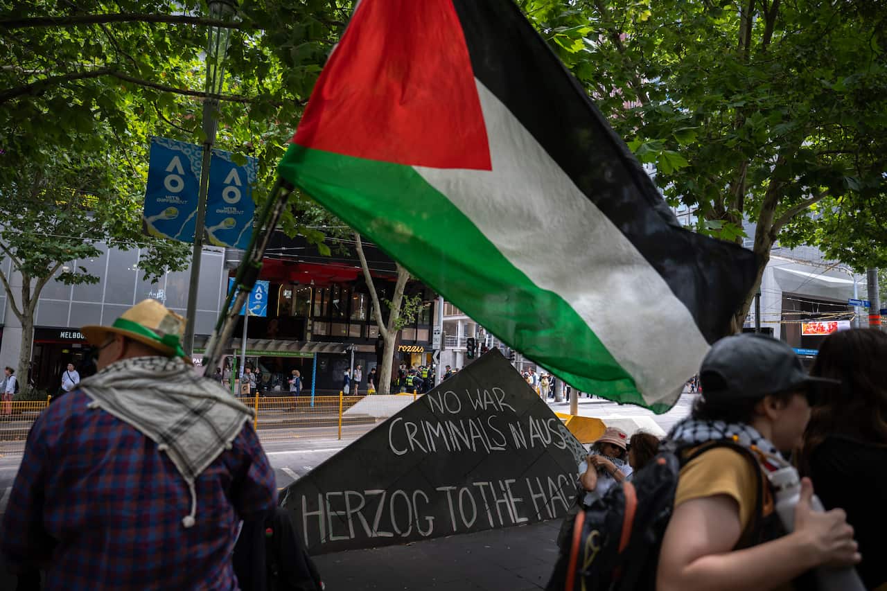 "No War Criminals in Aus, Herzog to the Hague" written in chalk behind a man holding the Palestinian flag during a 'Free Palestine' rally protesting the invitation of Israeli president Isaac Herzog to Australia.