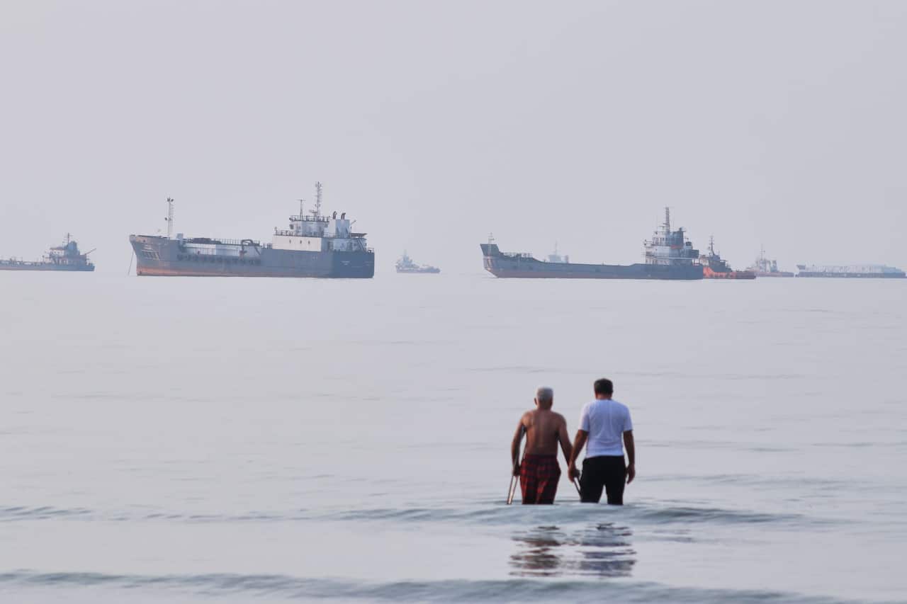 Two men wade in the waters of the Strait of Hormuz with vessels anchored in the background, off Bandar Abbas, Iran