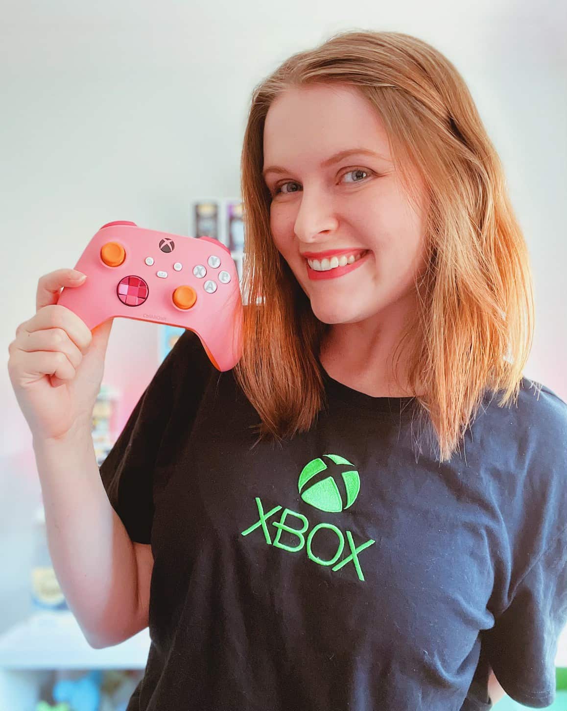 A woman holding a pink gaming controller, wearing a t-shirt with the green Xbox logo on it