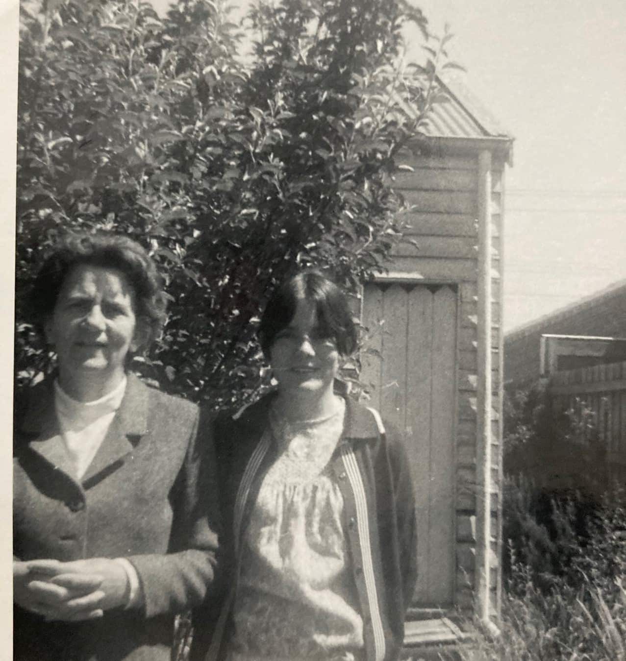 Black and white photograph of two women standing together. There is a tree and large wooden shed behind them.