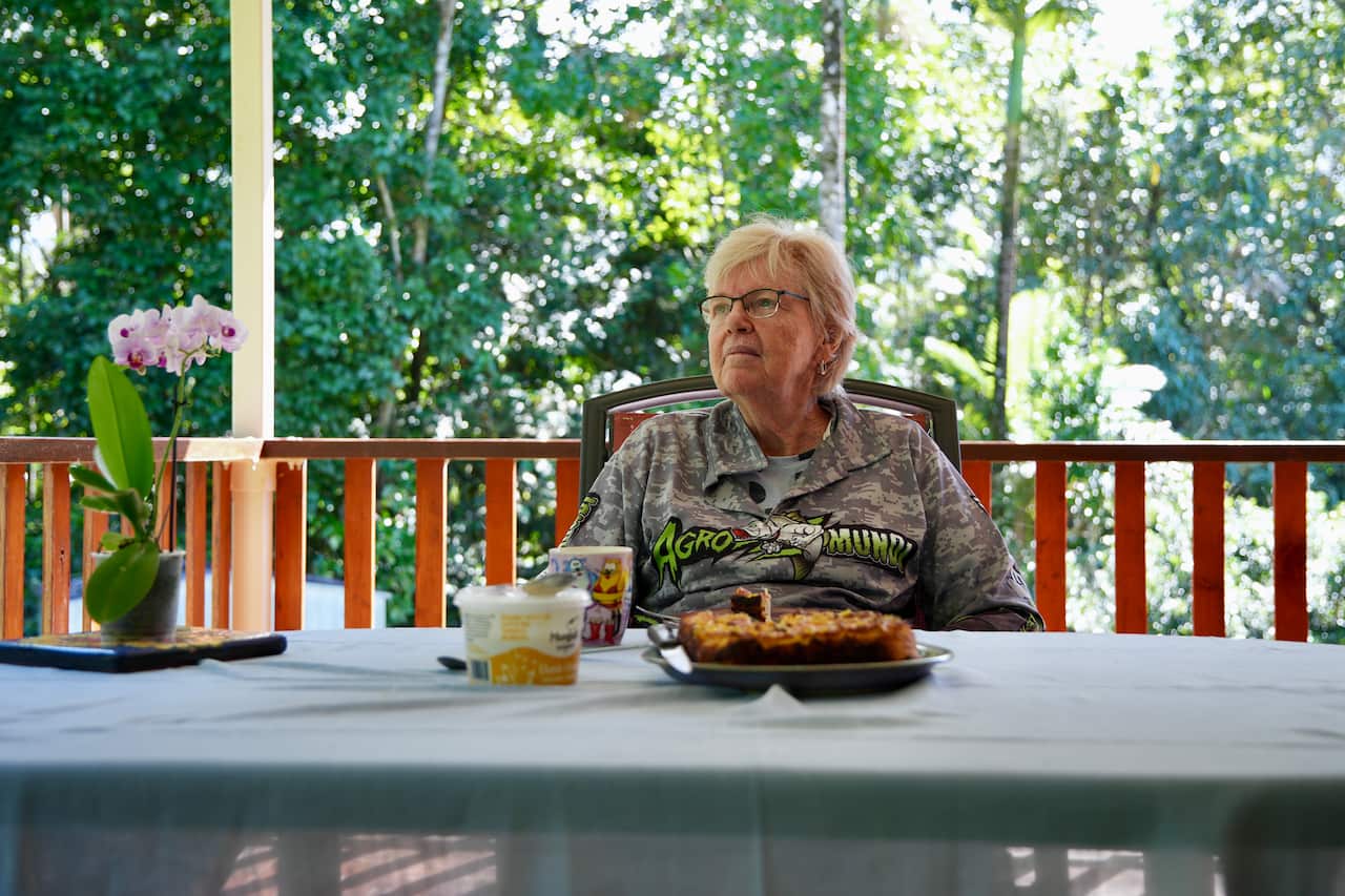 An older woman in a print-patterned t-shirt and glasses looks away as she sits at a table on a verandah. The table has a mug, a cake and a tub of butter on it. There are rainforest trees in the background