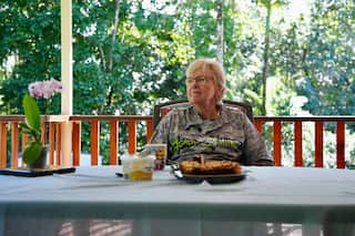 An older woman in a print-patterned t-shirt and glasses looks away as she sits at a table on a verandah. The table has a mug, a cake and a tub of butter on it. There are rainforest trees in the background