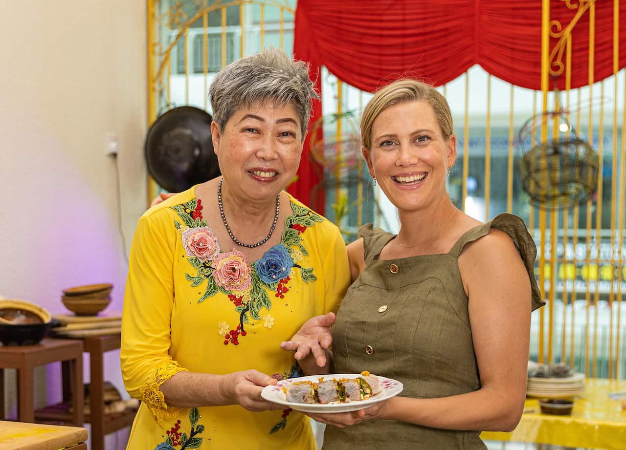 A woman in a vibrant yellow top stands beside another woman in a kitchen. Both are smiling. They are holding a plate between them. The plate has a filled roll on it, cut into pieces. 