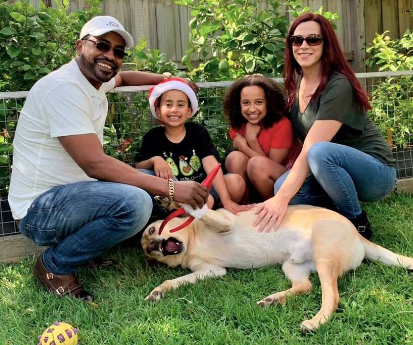 A man in a white shirt and cap crouches beside two children and a dog while a woman leans forward.