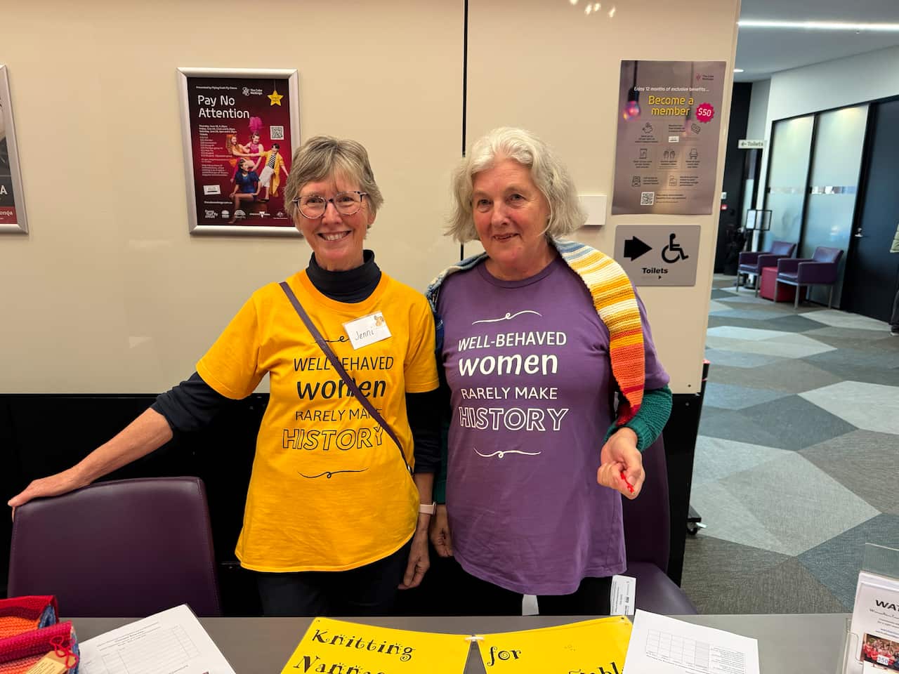 A woman in a yellow t-shirt stands next to a woman in a purple t-shirt inside a room.