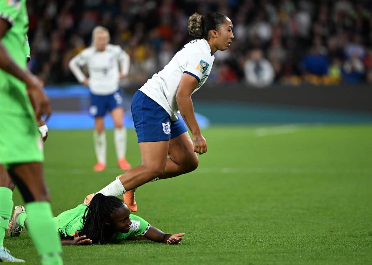 A woman in a white football uniform puts her foot on the back of a woman in a green football uniform