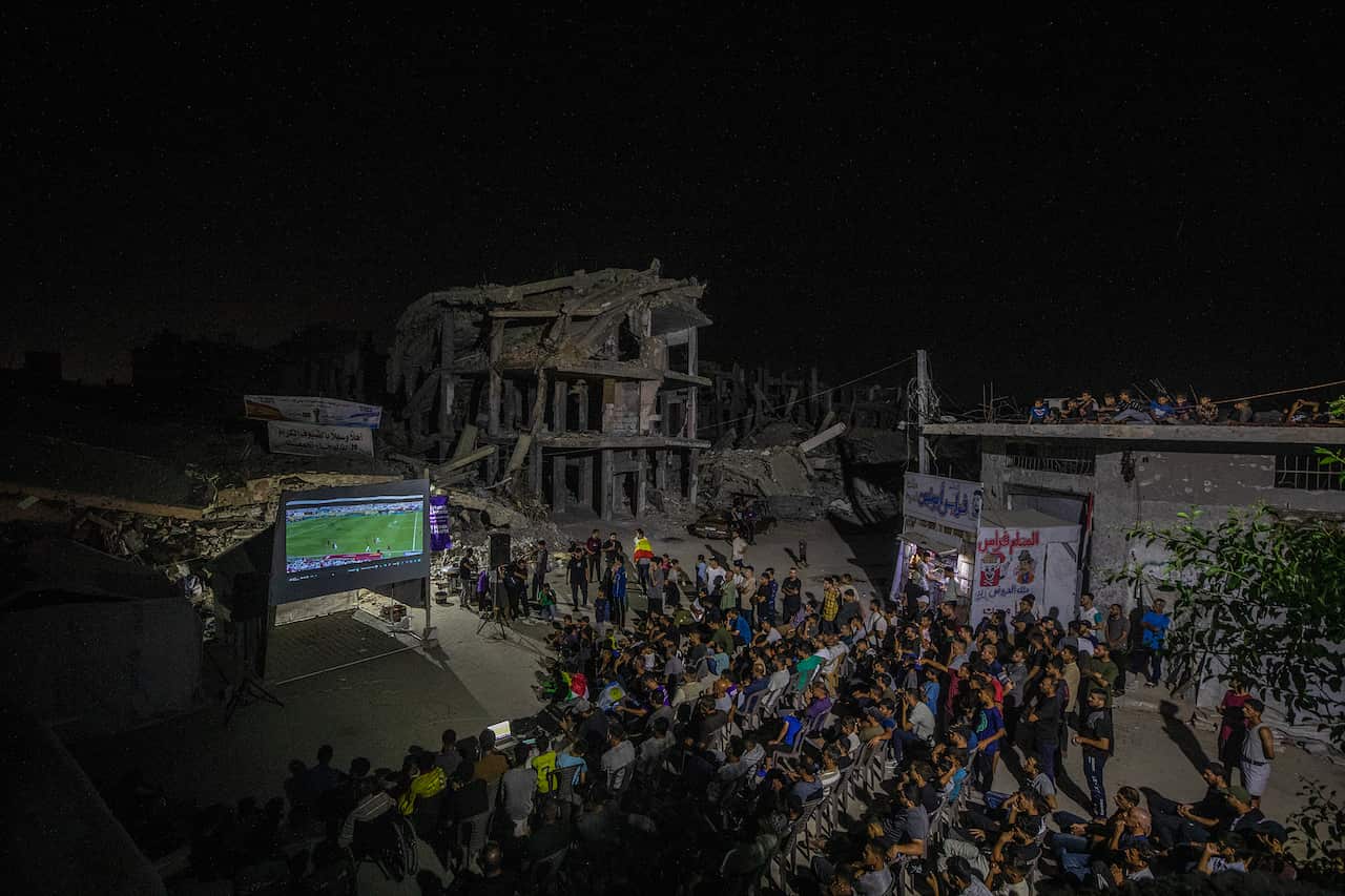 A crowd of people watches a football match on a projector in a heavily damaged street under cover of darkness.