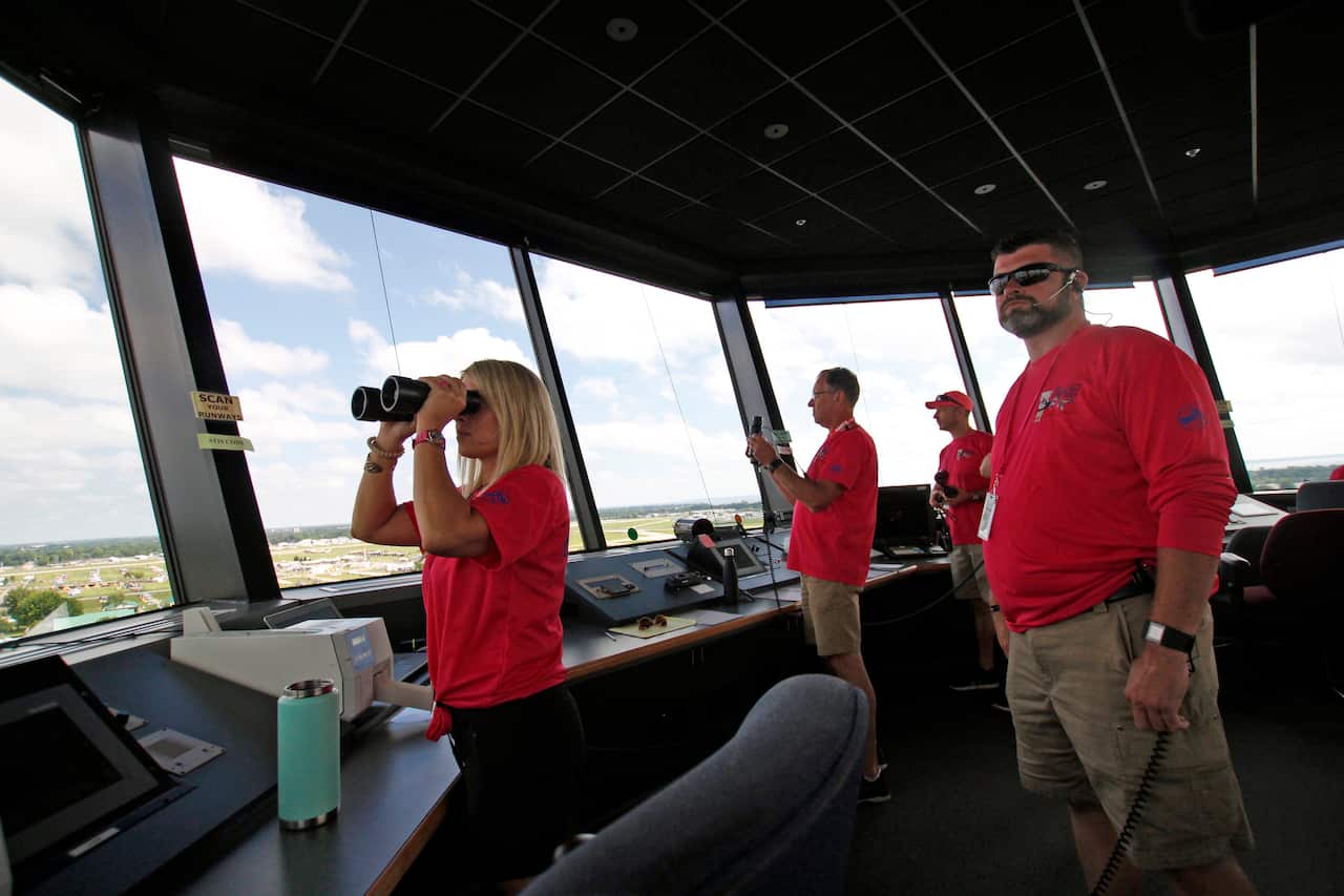 Air traffic controllers wearing bright red tops stare outside a large window in an air traffic control tower. A woman with blonde hair holds binoculars to her face.