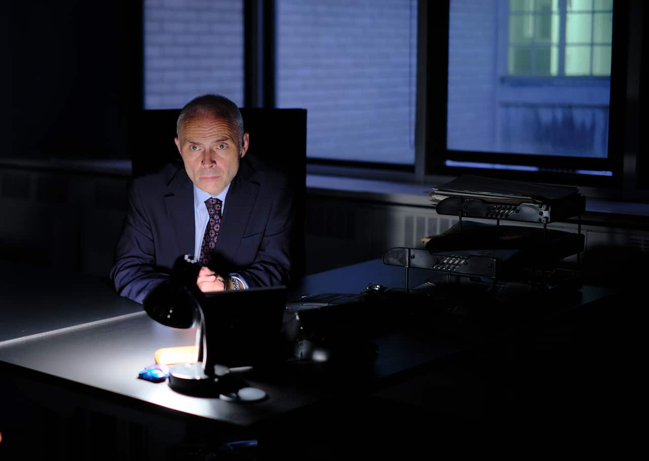 A man in a suit sits in a dark office, with the light of a desk lamp reflecting on his face. 