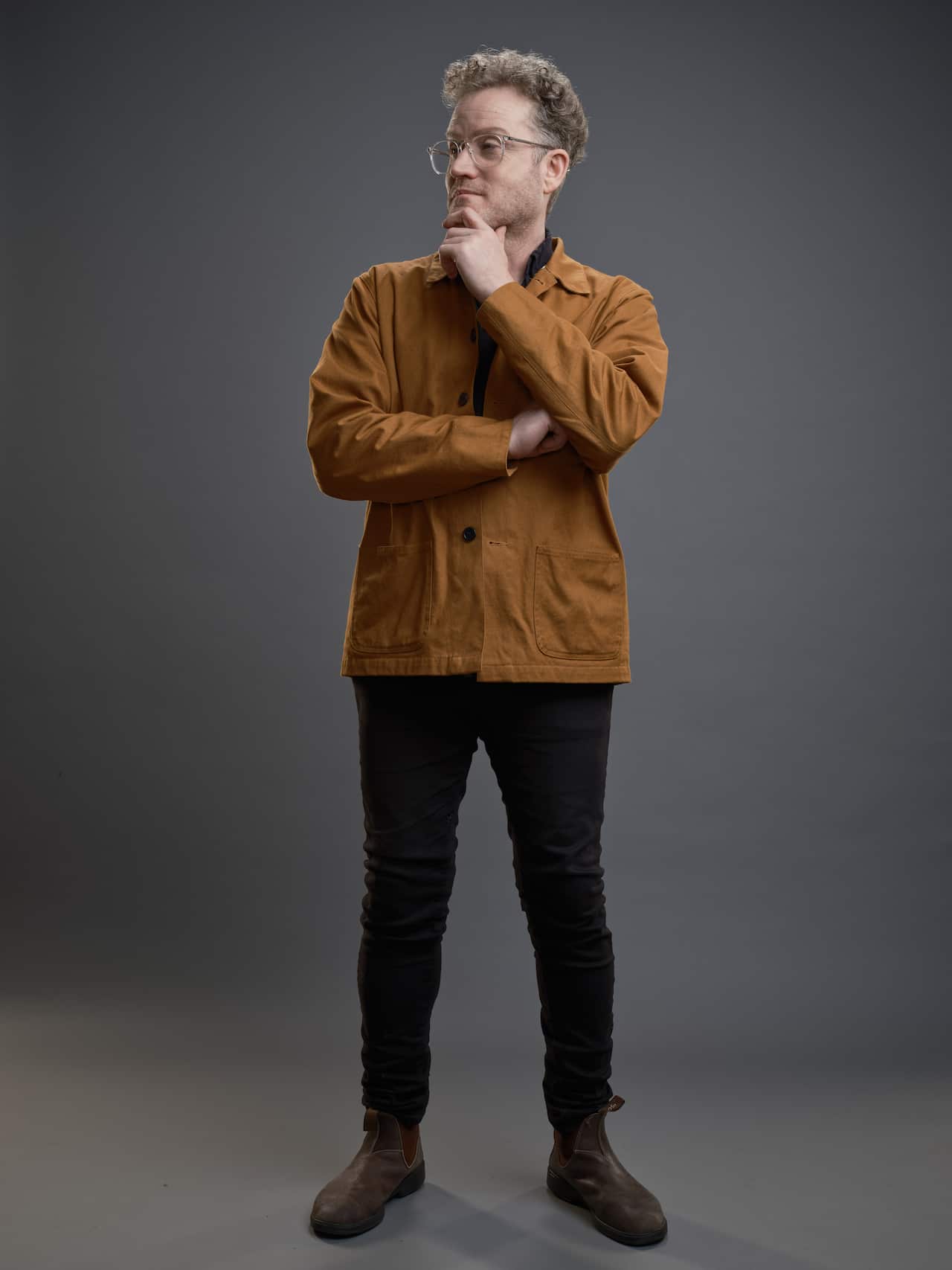 A man stands in a pose, wearing black pants and a brown shirt. 
