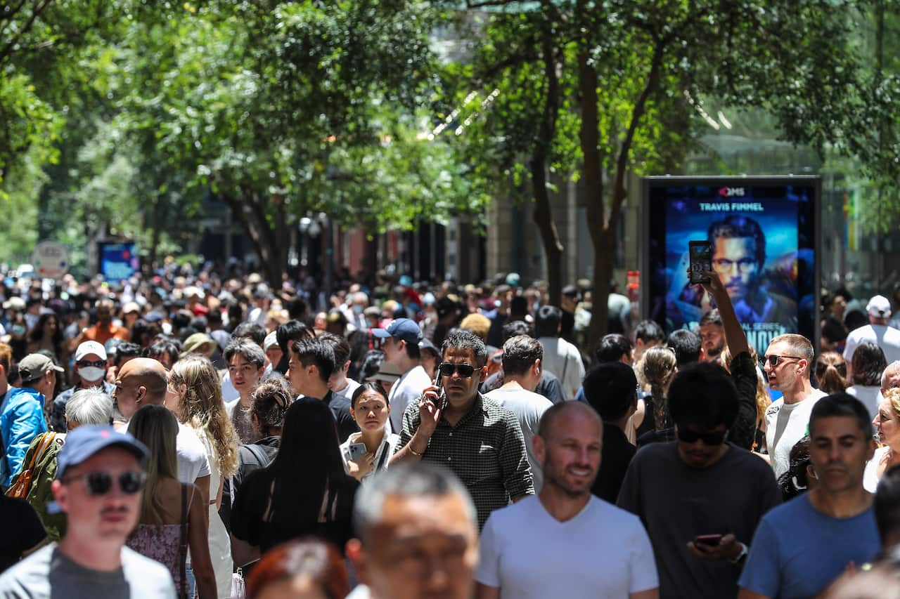 A busy crowd of people from various demographics walk the streets of Sydney, with shops and lush trees in the background.