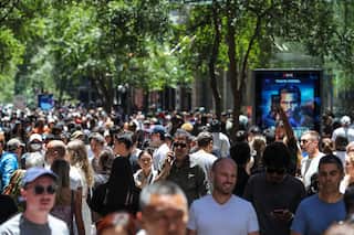 A busy crowd of people from various demographics walk the streets of Sydney, with shops and lush trees in the background.
