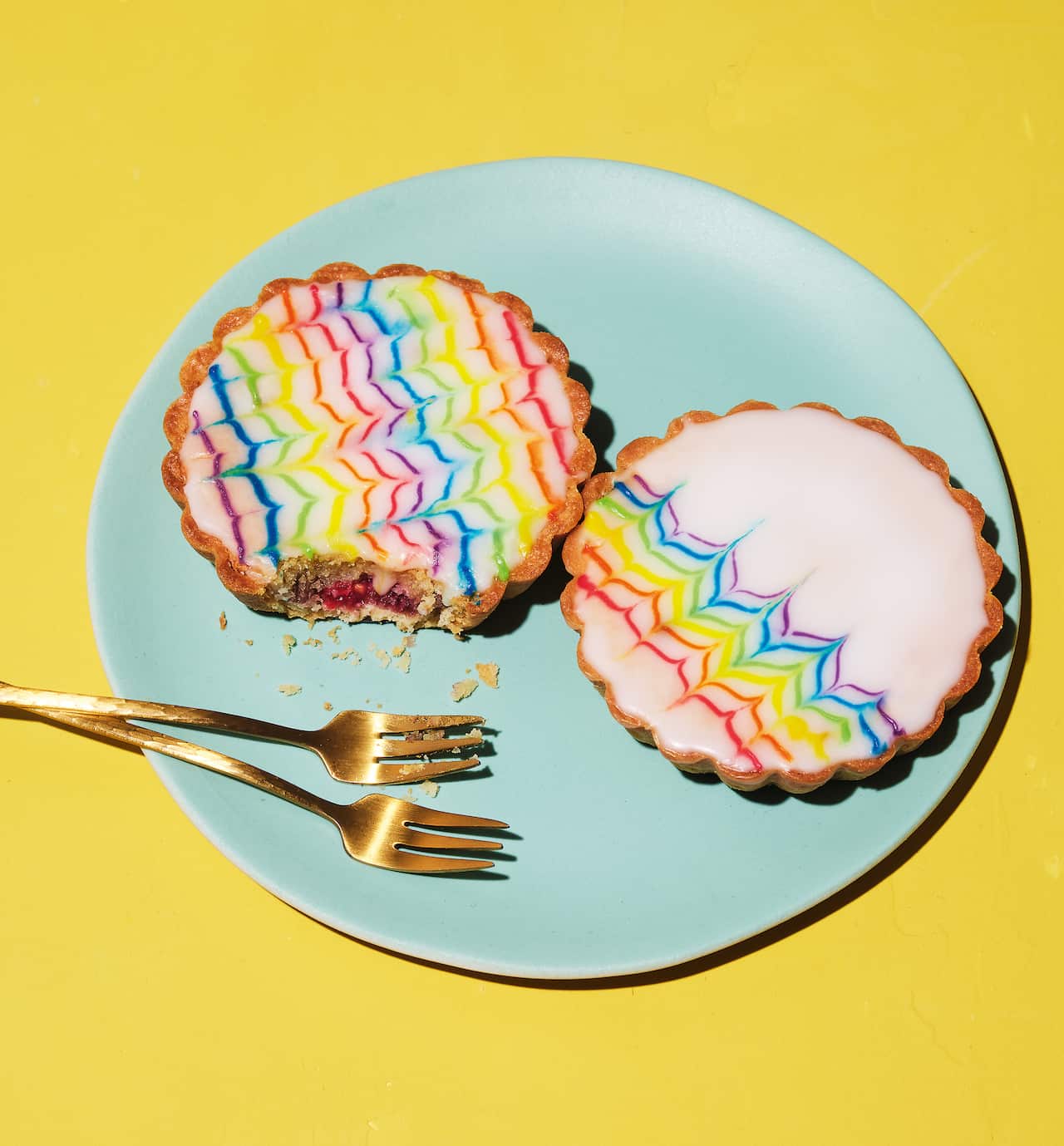 Two small round tartletes with fluted pastry edges sit on a blue plate on a bright yellow background. Each tart is decorated with white icing with a feathered pattern in rainbow colours.