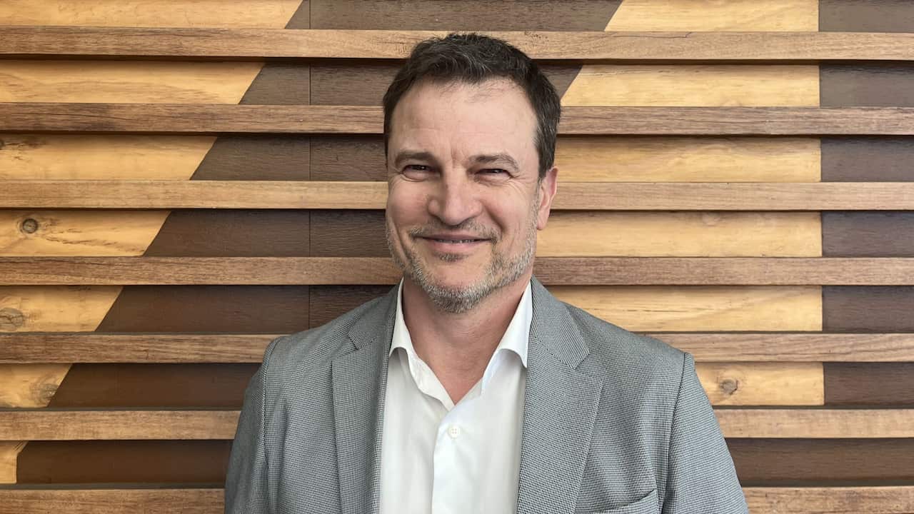 a middle aged man in a grey suit smiles in front of a timber background