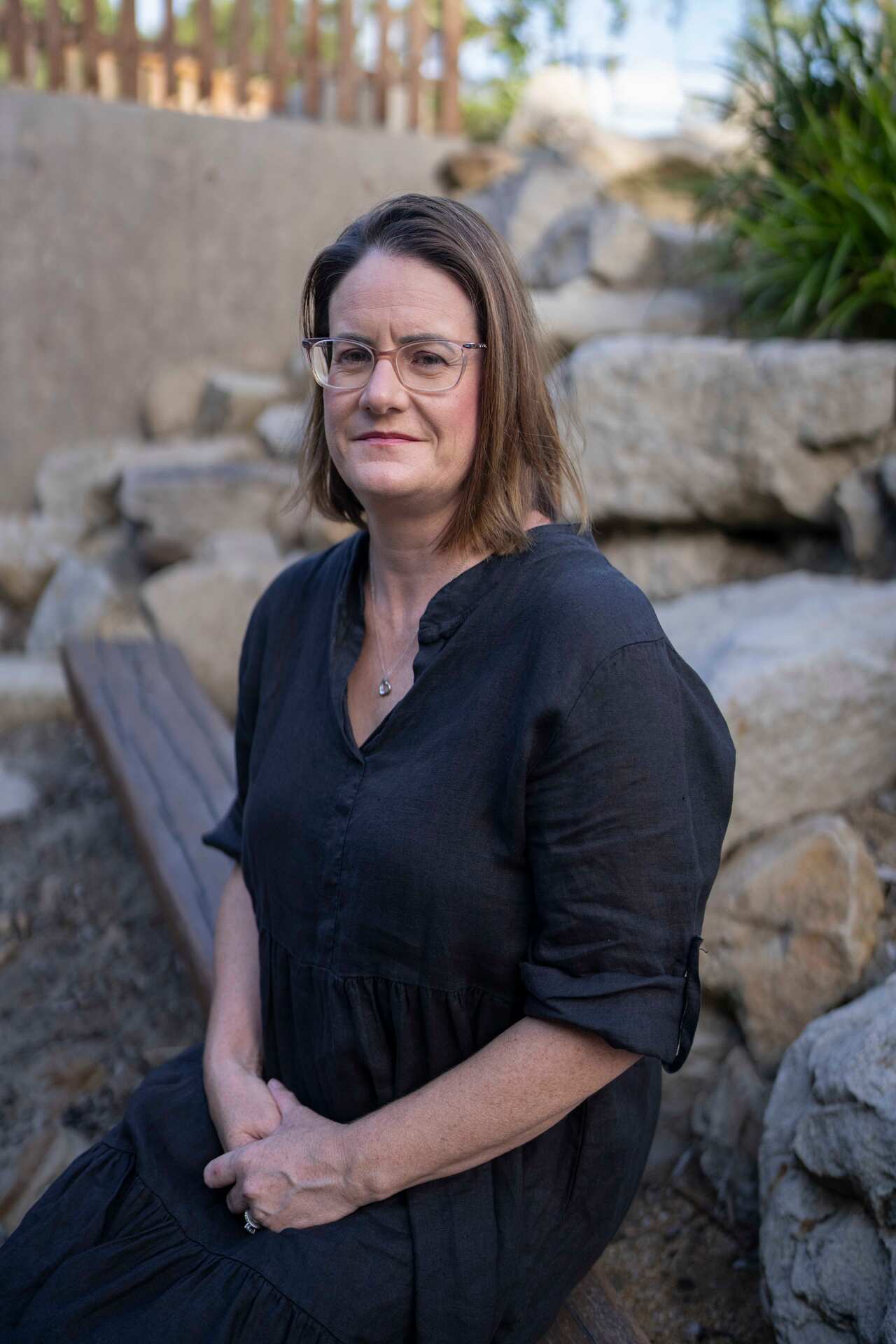 A middle-aged woman with short dark hair and wearing a black dress sits on a wooden bench in a park.