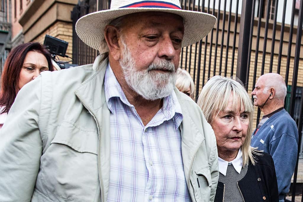 A man with a grey beard and a hat walks with a woman with shoulder-length blonde hair. They both have solemn expressions.