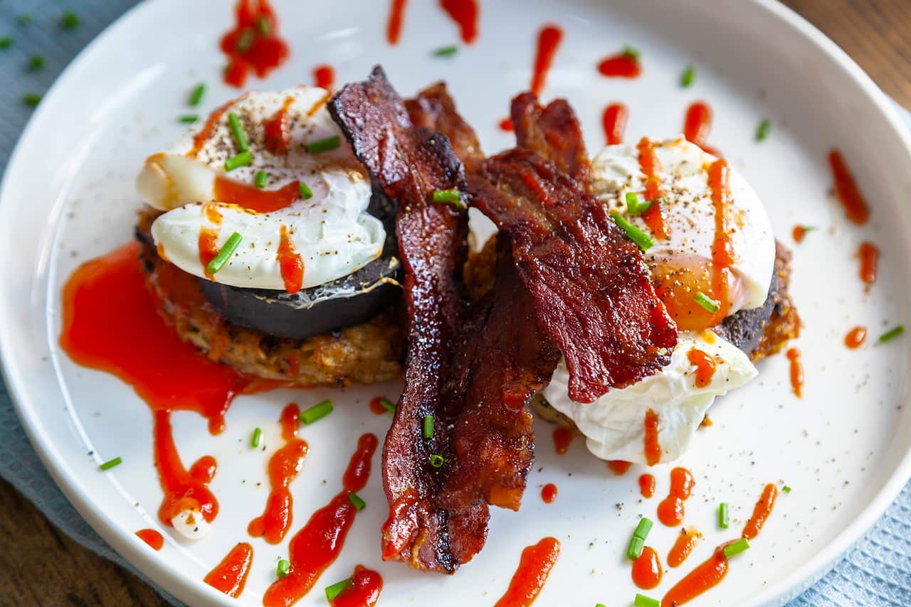 Two stacks, each with several layers including a rosti pattie and an egg, sit side by side on a white plate, drizzled wiht a reddish sauce. Several pieces of crispy bacon sit on top. 
