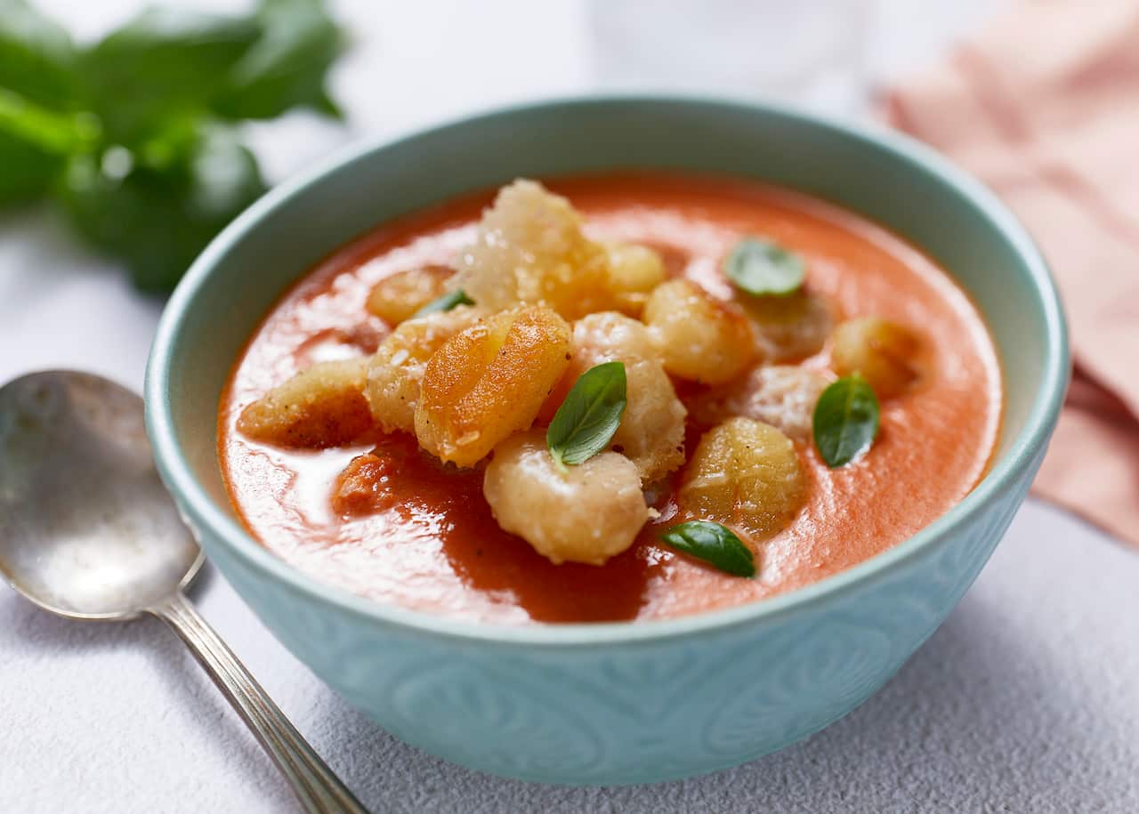 A blue soup bowl with a patterned outside surface holds tomato soup topped with a tumble of cooked gnocchi and small basil leaves. A burnished silver soup spoon sits alongside. 