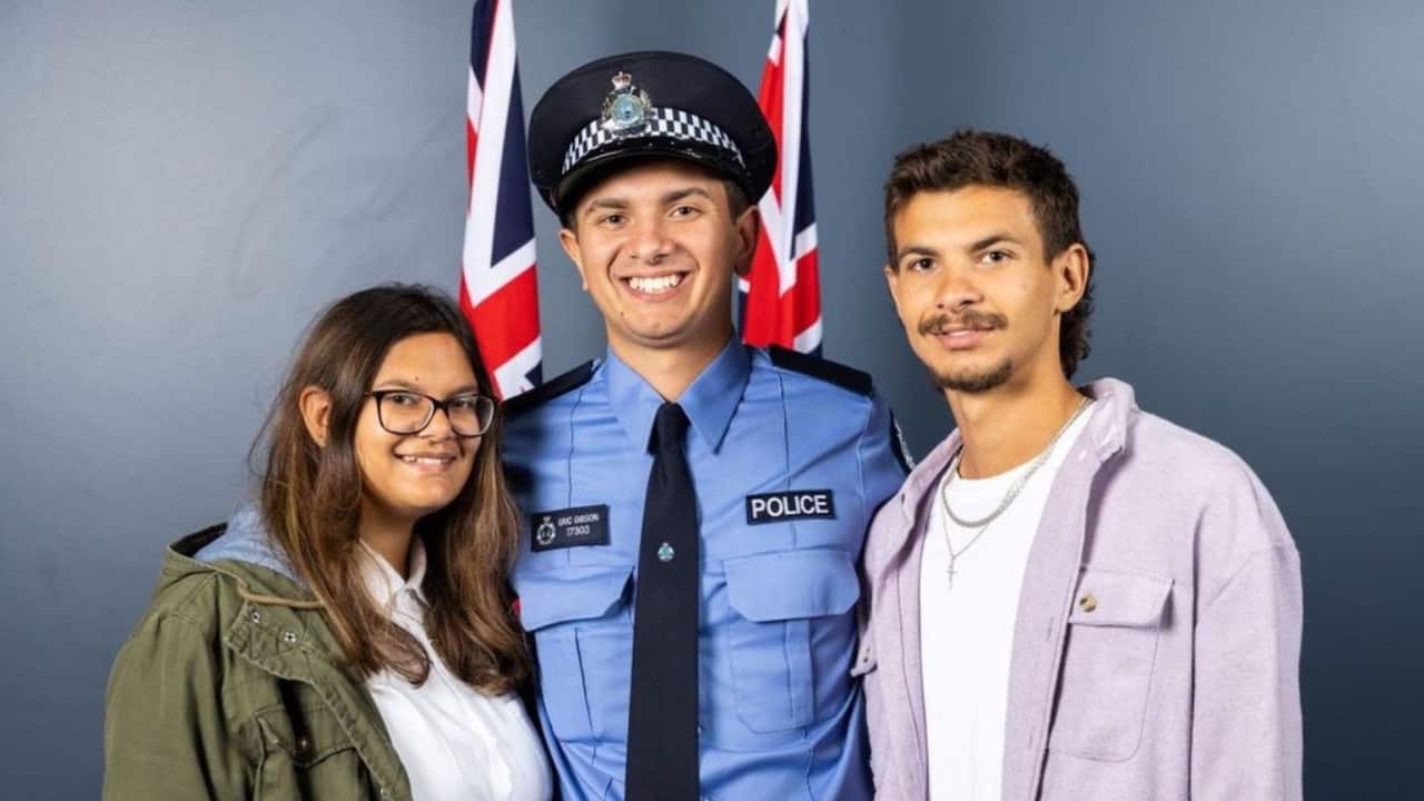 A young man wearing a blue police uniform with hat and tie stands between a young man and woman inside. There are two Australian flags behind them.
