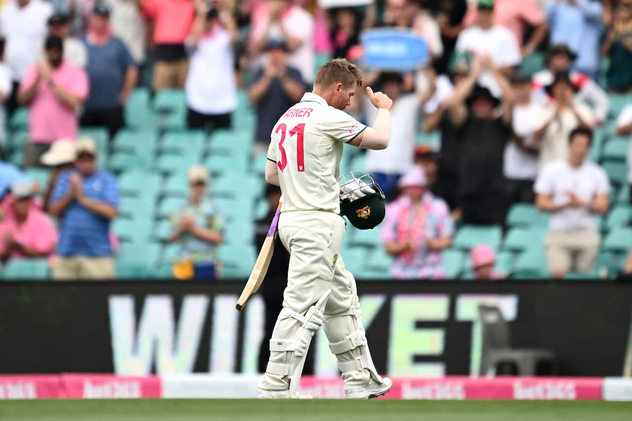 A man in a cricket uniform holding a cricketer's helmet walks across a cricket field.