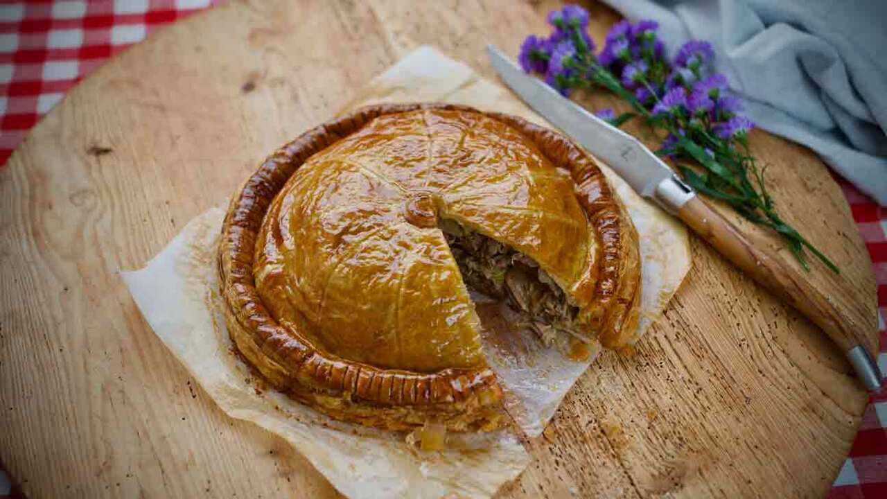A round pie with golden pastry and a slice cut out to show the meat filling sits on a wooden surface. 