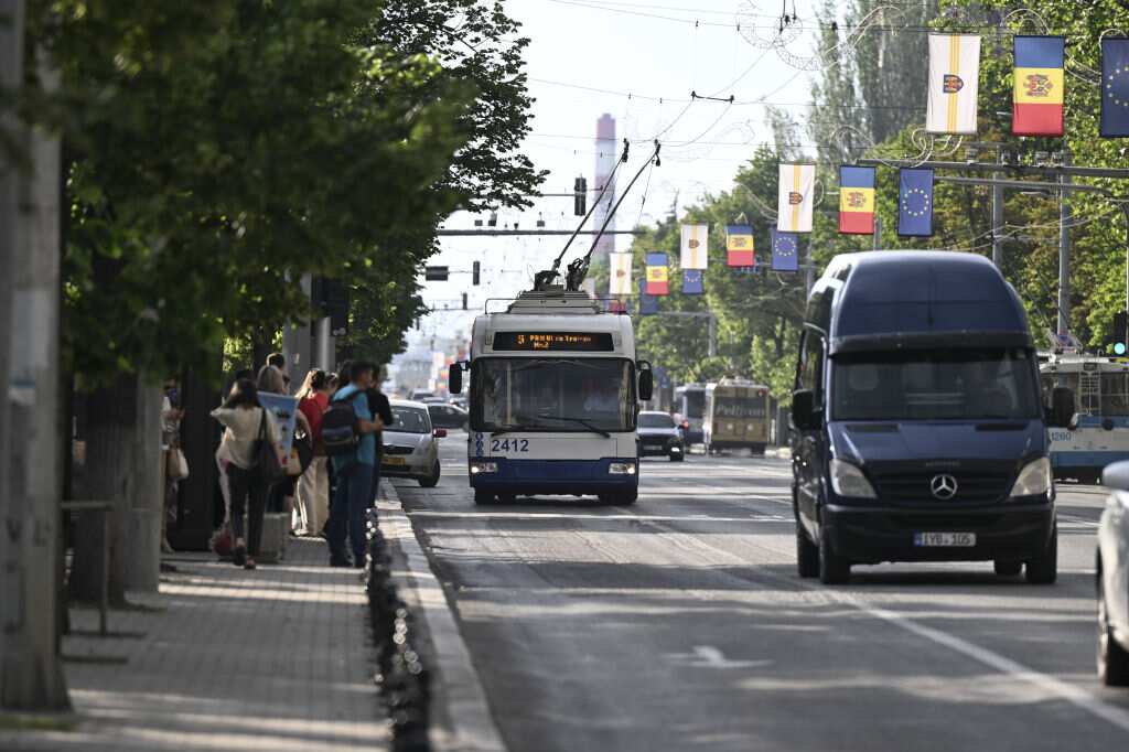 A trolley bus and van drive along a busy road. Pedestrians walk on a footpath beside the road. 