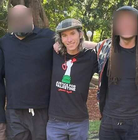 A thin man in a vintage-style flat cap stands with arms around the shoulders of two other men. 