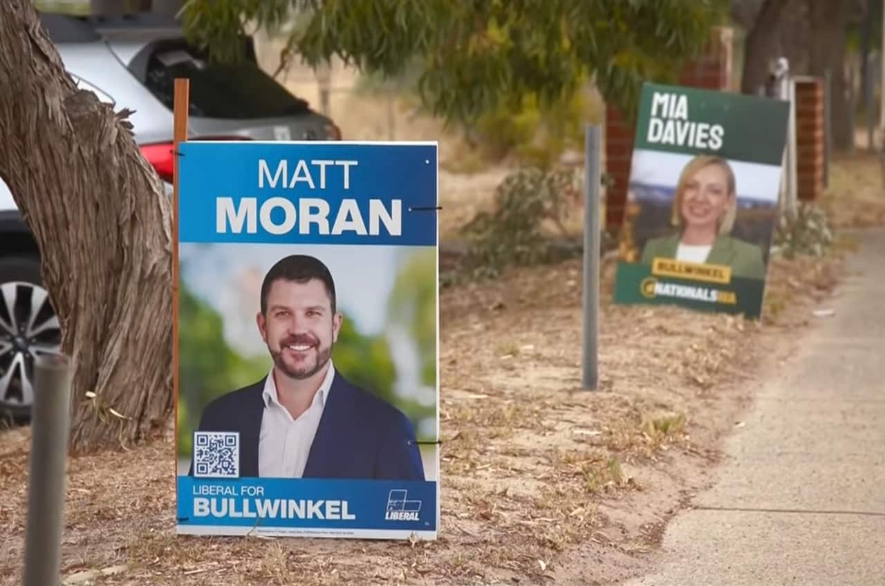 Two election campaign signs one after the other, on a roadside. One is blue and reads Matt Moran above a picture of a man. The other is green and reads Mia Davies above a picture of a woman.