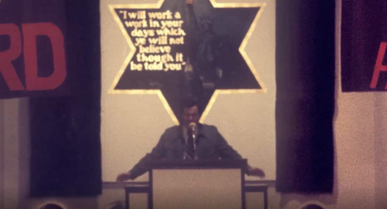 An old image of a man preaching behind a lectern and microphone. Behind him is a six-pointed star with text in it reading: "I will work a work in your days which ye will not believe though it be told you."